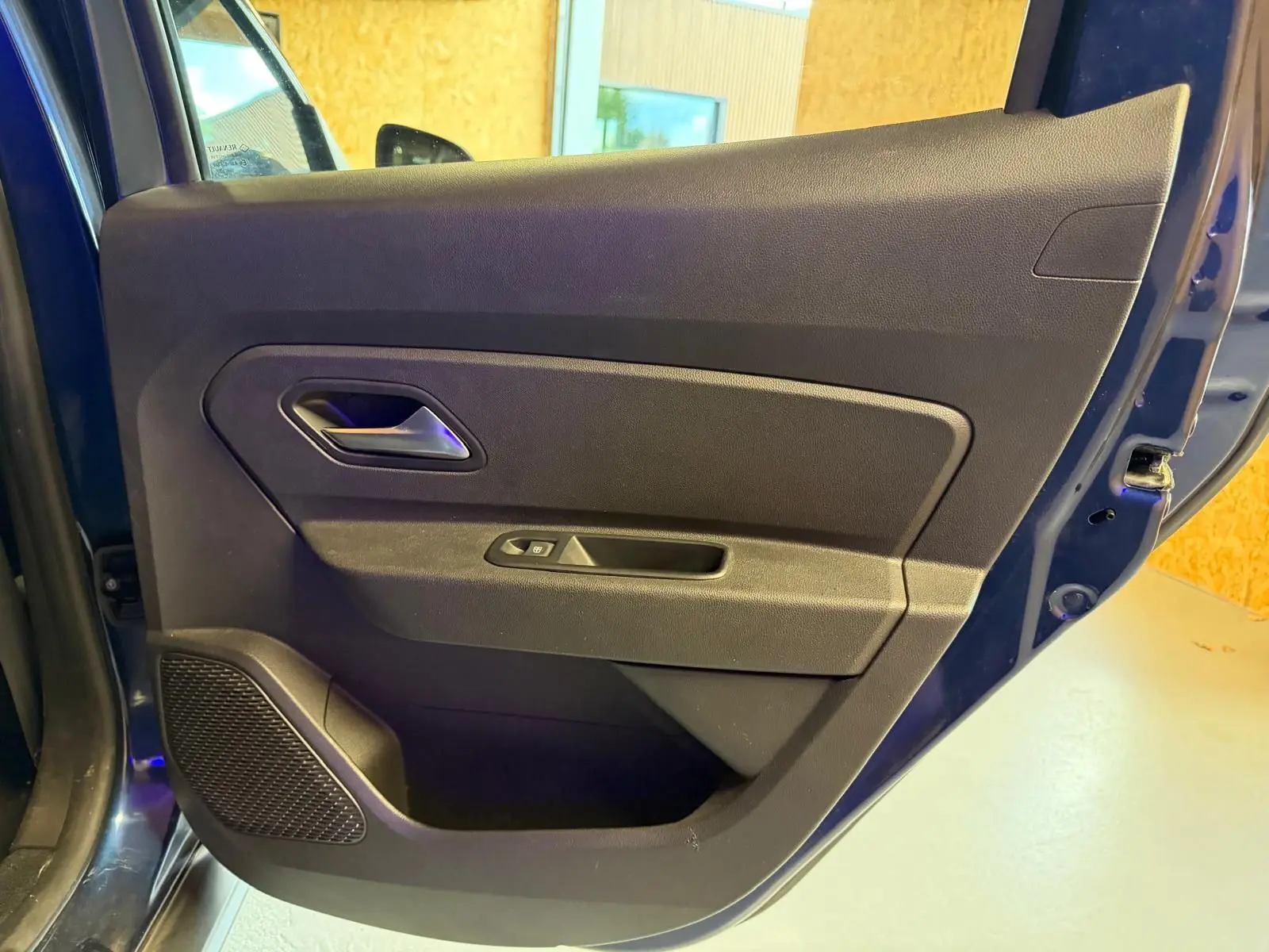 Vue côté gauche de la porte avant bleue du Dacia Duster, avec poignée intérieure et commande de vitre électrique.