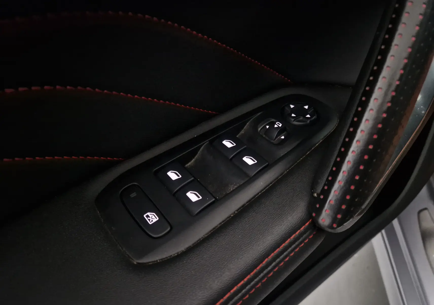 Gros plan sur les commandes de lève-vitres et rétroviseurs côté conducteur, cuir noir avec surpiqûres rouges.