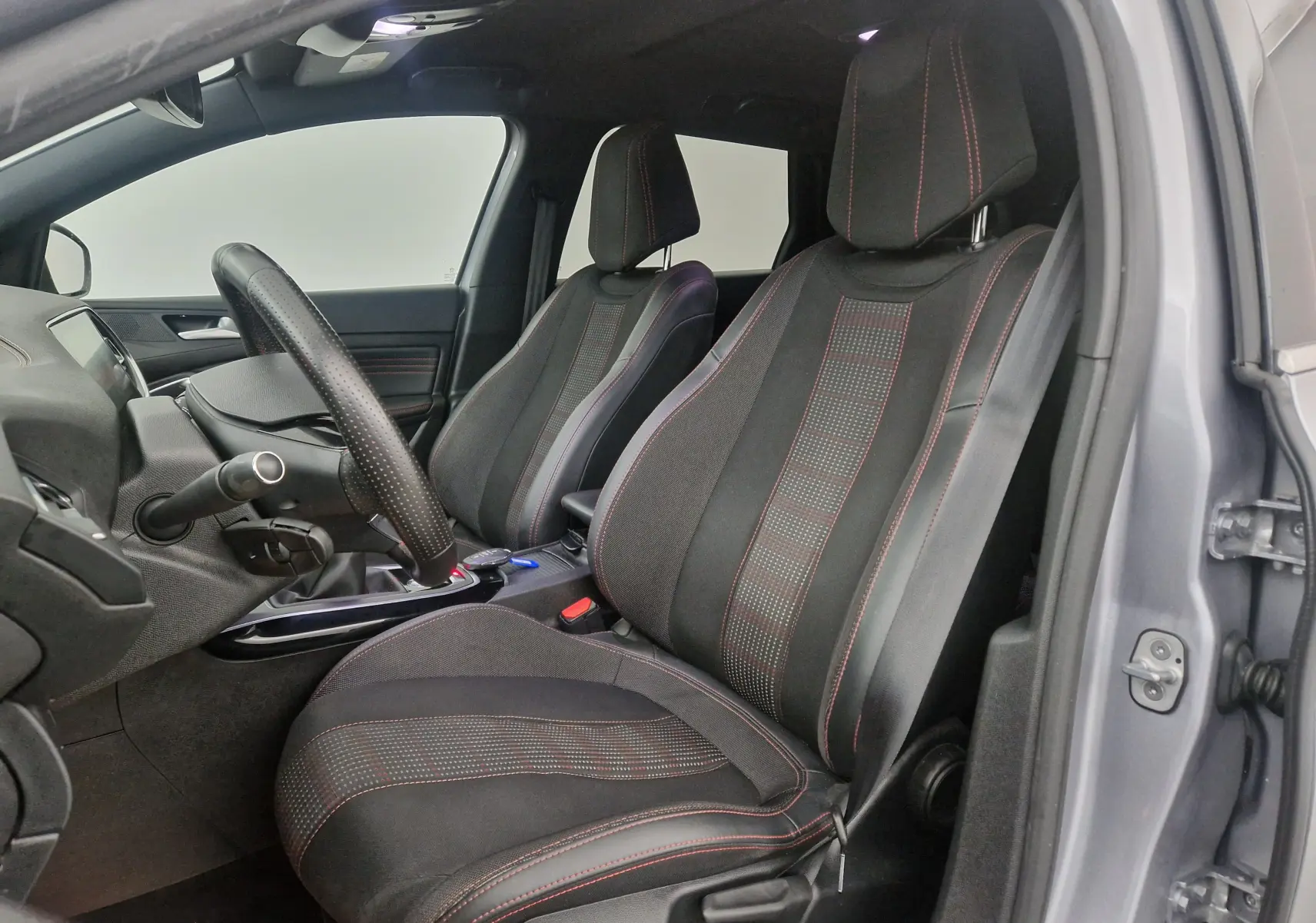 Vue intérieure côté conducteur de la Peugeot 308 SW gris Artense, sièges sport noirs avec surpiqûres rouges et volant cuir perforé.