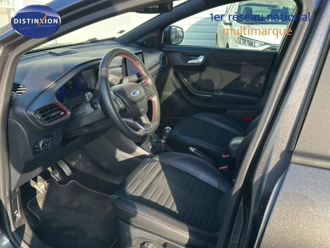 Vue intérieure côté conducteur d'un Ford Puma gris foncé, montrant sièges noirs avec surpiqûres rouges et tableau de bord moderne.