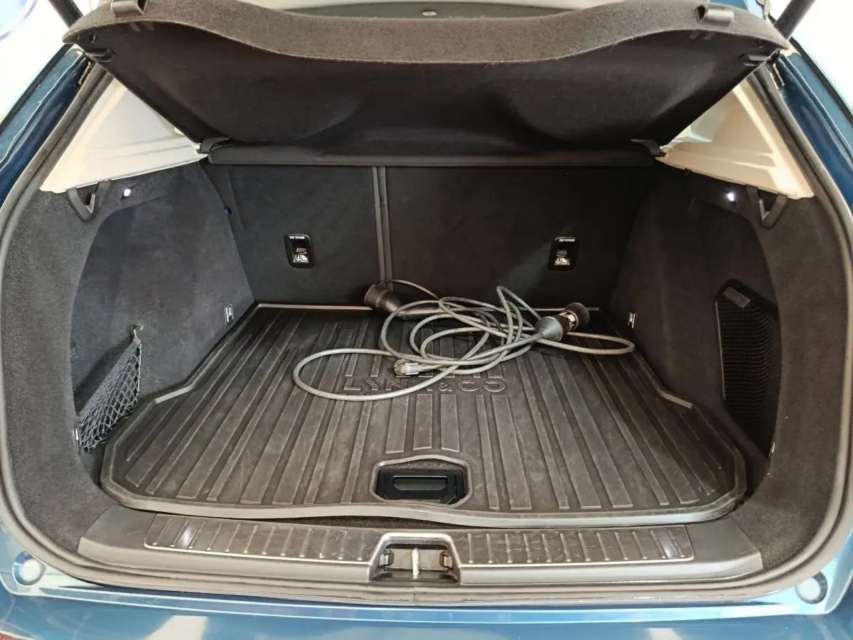 Coffre ouvert vu de l'arrière d'un Link & Co 01 bleu avec câble de recharge hybride posé sur le tapis de coffre.