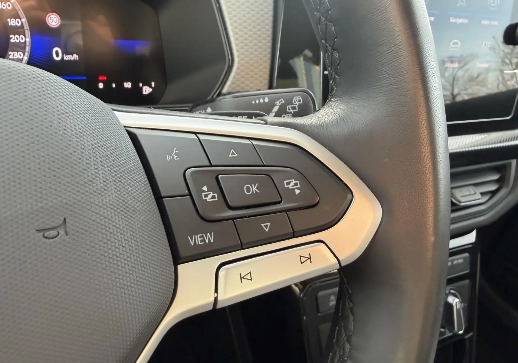 Gros plan sur les commandes multifonctions du volant cuir noir du Volkswagen T-Cross gris, avec écran digital en arrière-plan.