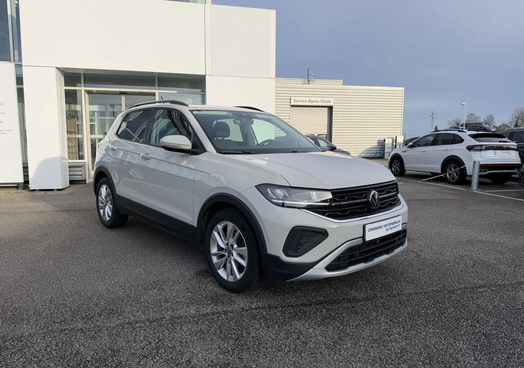Volkswagen T-Cross gris en 3/4 avant droit, avec jantes alliage et calandre noire, stationné devant un bâtiment.