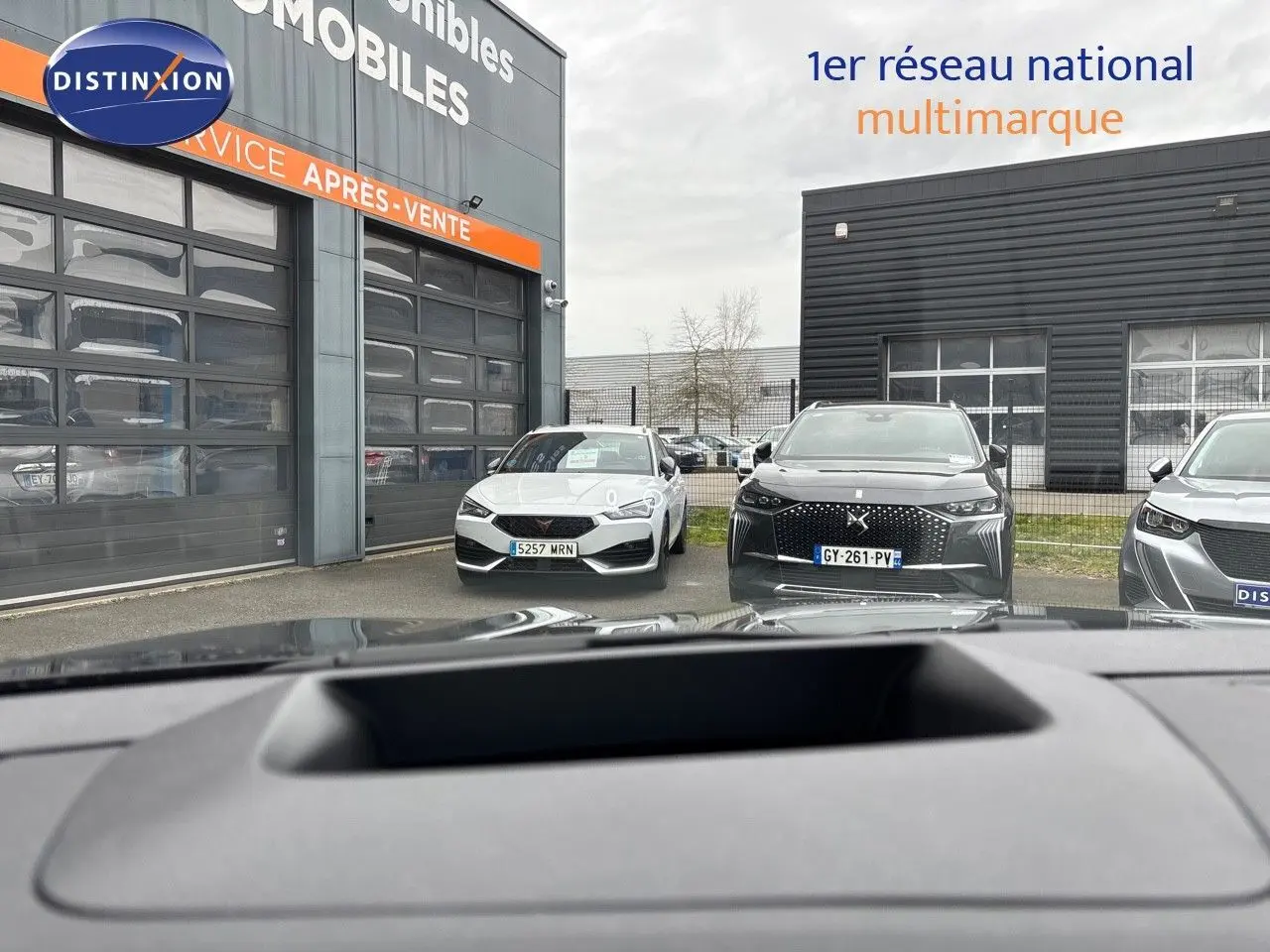 Vue depuis l'intérieur montrant le tableau de bord et par la vitre avant un Renault Austral noir étoile métal en 3/4 avant.