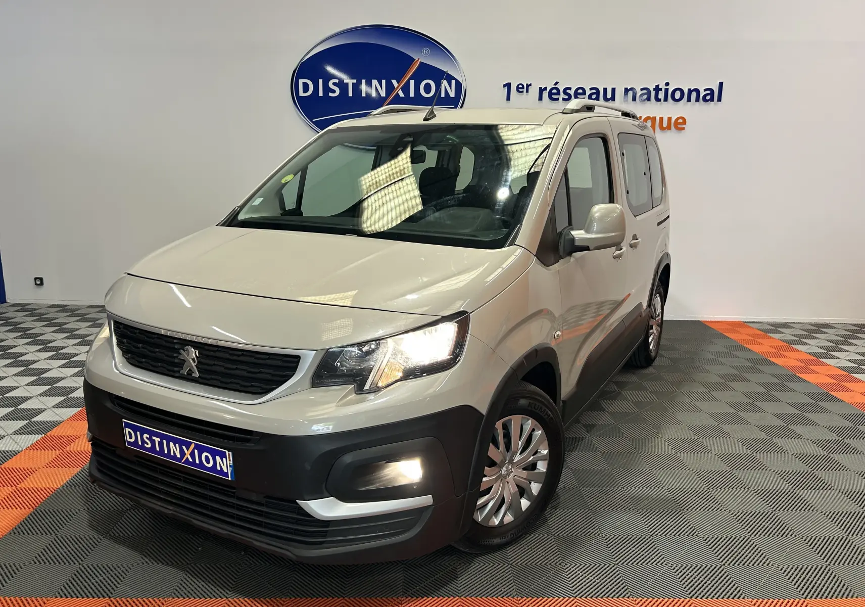 Peugeot Rifter gris sable en 3/4 avant droit, avec attelage et phares allumés dans un showroom.