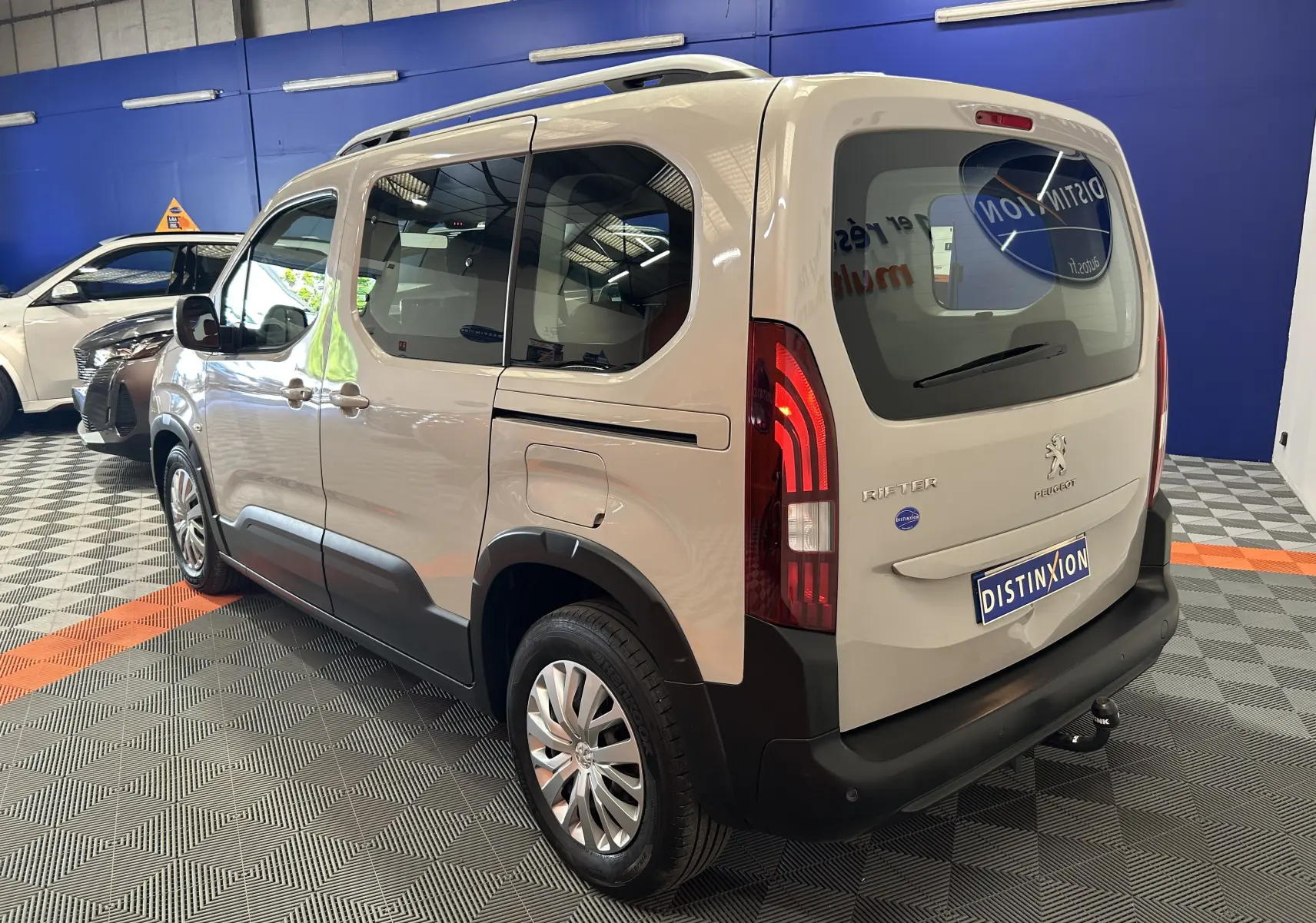 Vue 3/4 arrière droite du Peugeot Rifter gris sable 2019 avec attelage et vitres teintées en intérieur showroom.