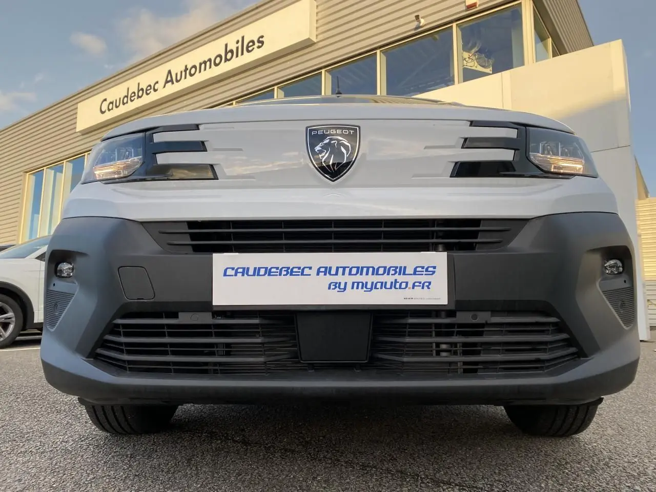Vue frontale basse du Peugeot Partner Fourgon blanc avec logo lion et calandre noire, devant un bâtiment commercial.