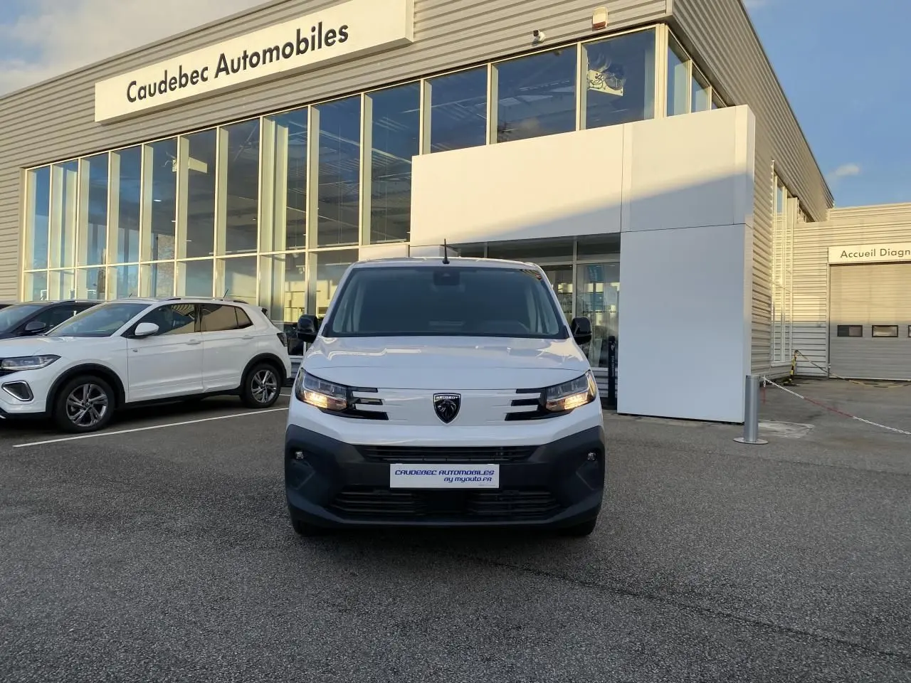 Vue de face du Peugeot Partner Fourgon blanc stationné devant un concessionnaire avec calandre noire et phares allumés.