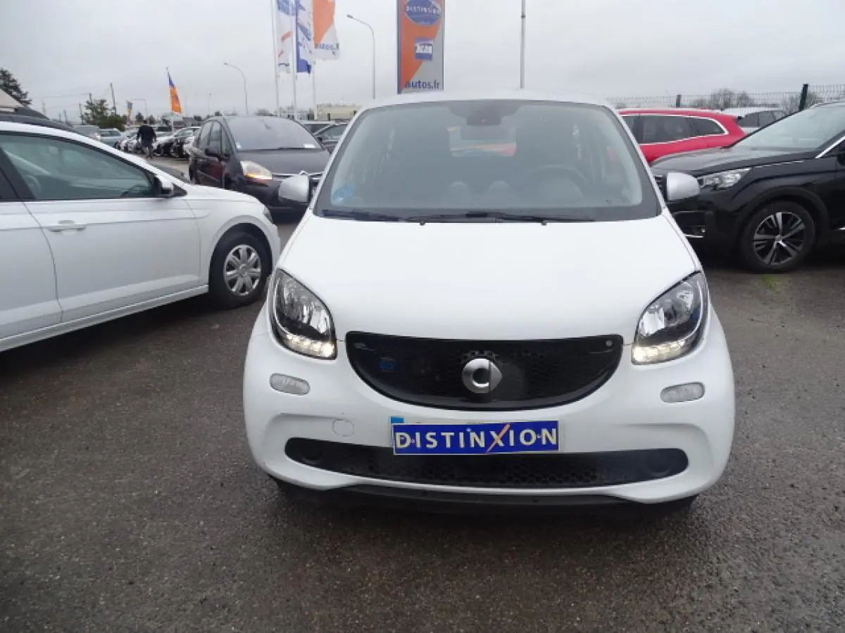 Vue de face d'une Smart Forfour électrique blanche avec calandre noir brillant et feux allumés sur parking.