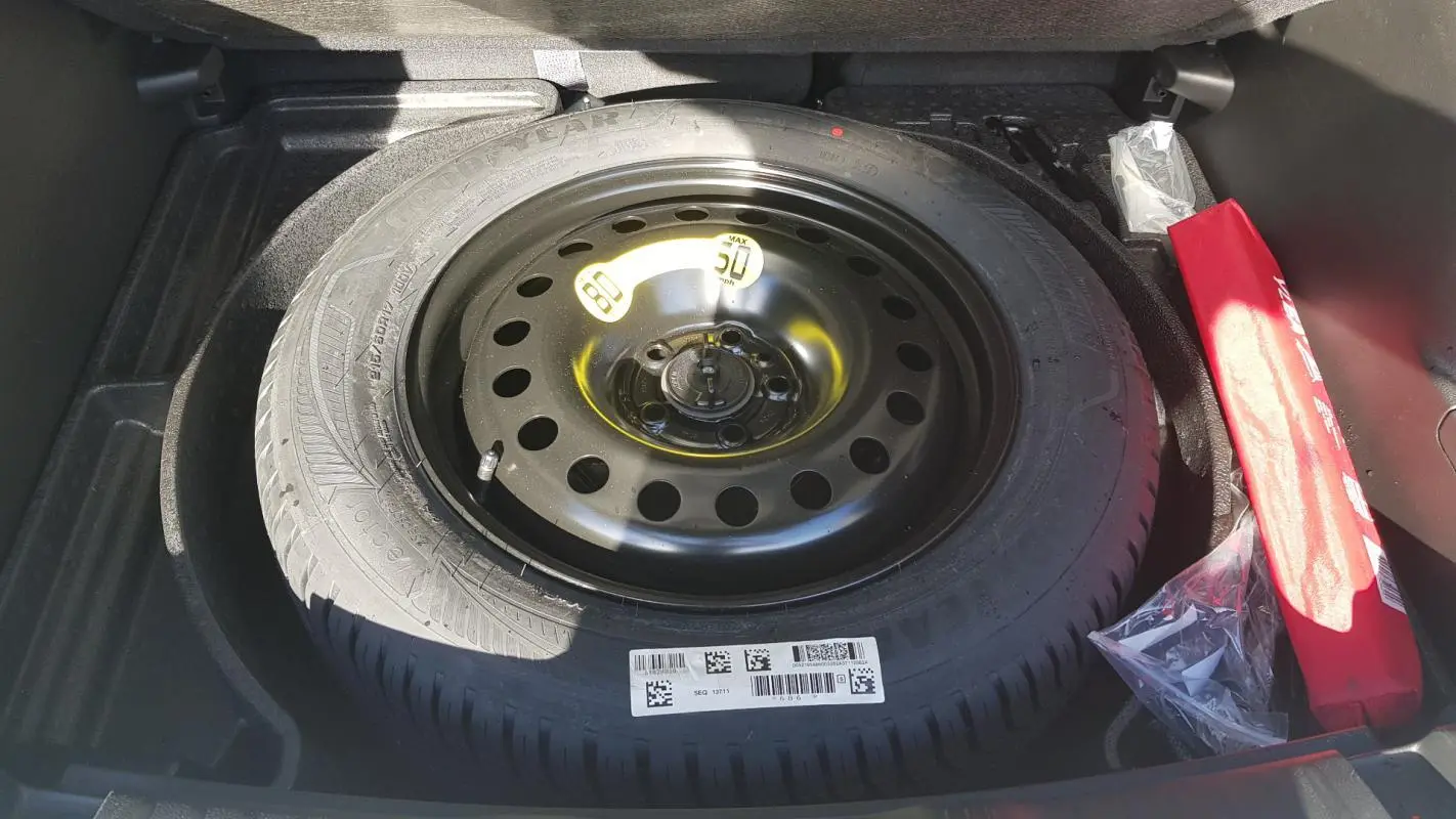 Roue de secours noire en acier dans le coffre du Jeep Compass blanc 2024, avec kit de réparation visible à droite.