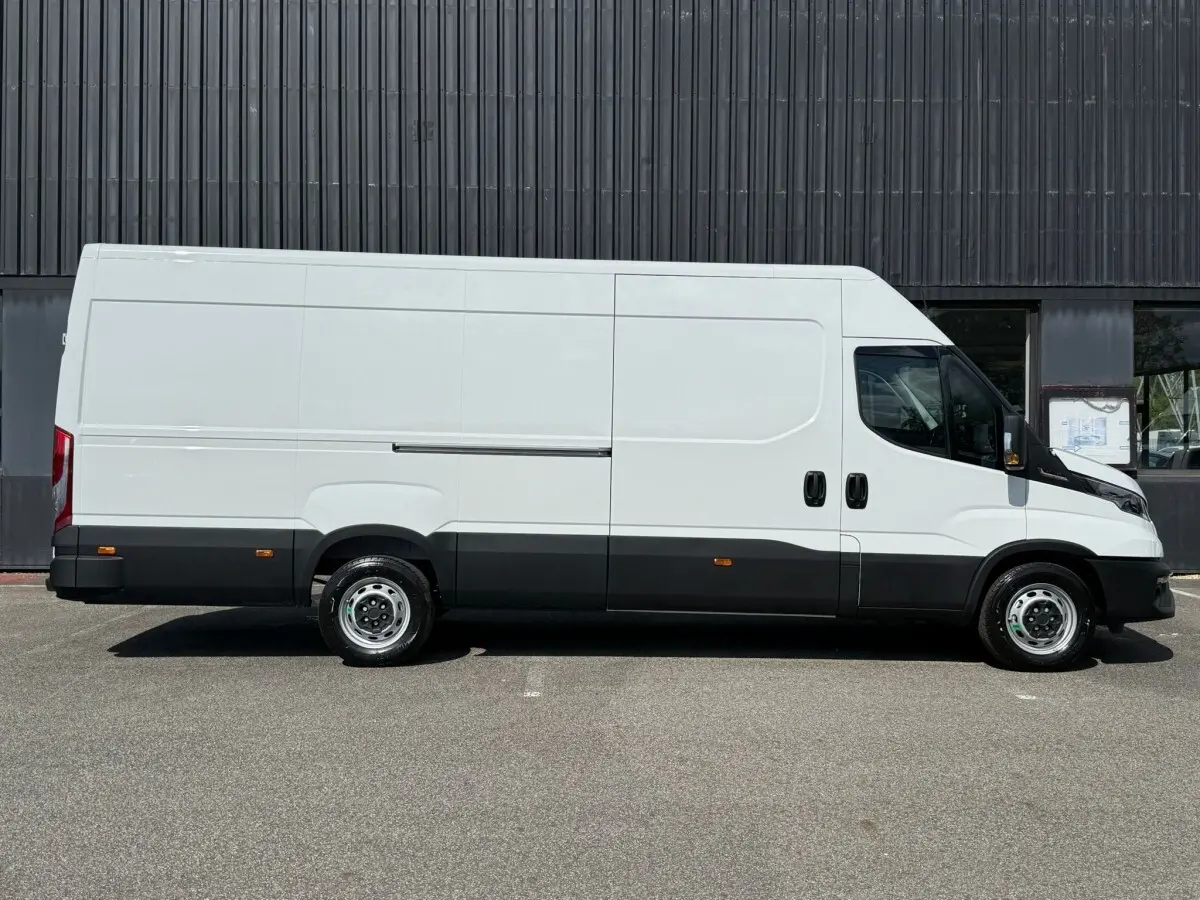 Fourgon Iveco Daily blanc vu de profil côté gauche, avec bas de caisse noir et roues acier, stationné devant un mur sombre.