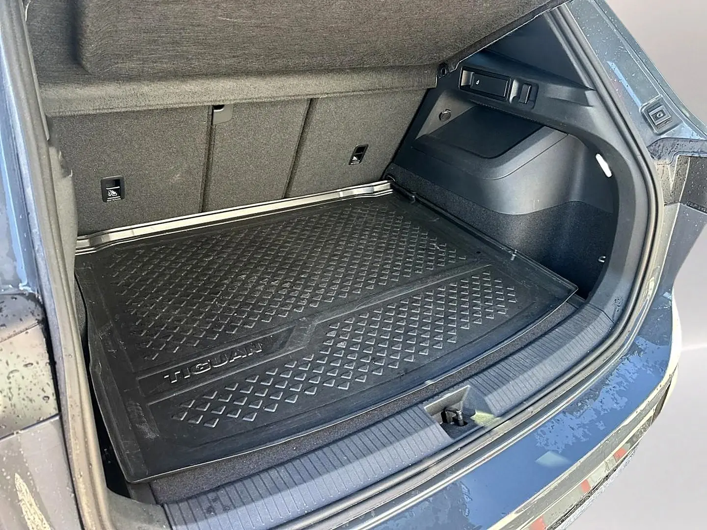Coffre ouvert vu de l'arrière droit d'un Volkswagen Tiguan gris dauphin avec tapis de protection noir marqué Tiguan.