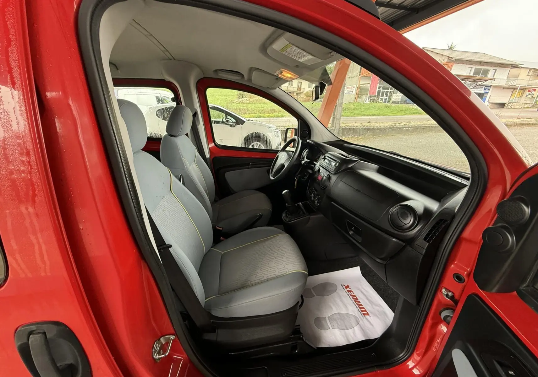Vue intérieure côté conducteur du Peugeot Bipper Tepee rouge, montrant sièges gris et tableau de bord noir.