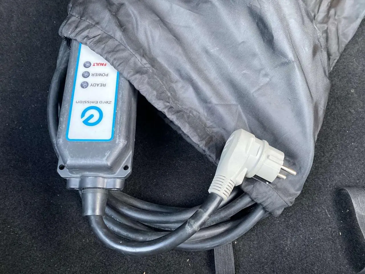 Gros plan sur le câble de recharge électrique noir avec boîtier blanc marqué "Zero Emission" dans le coffre d'une Nissan Leaf blanche.