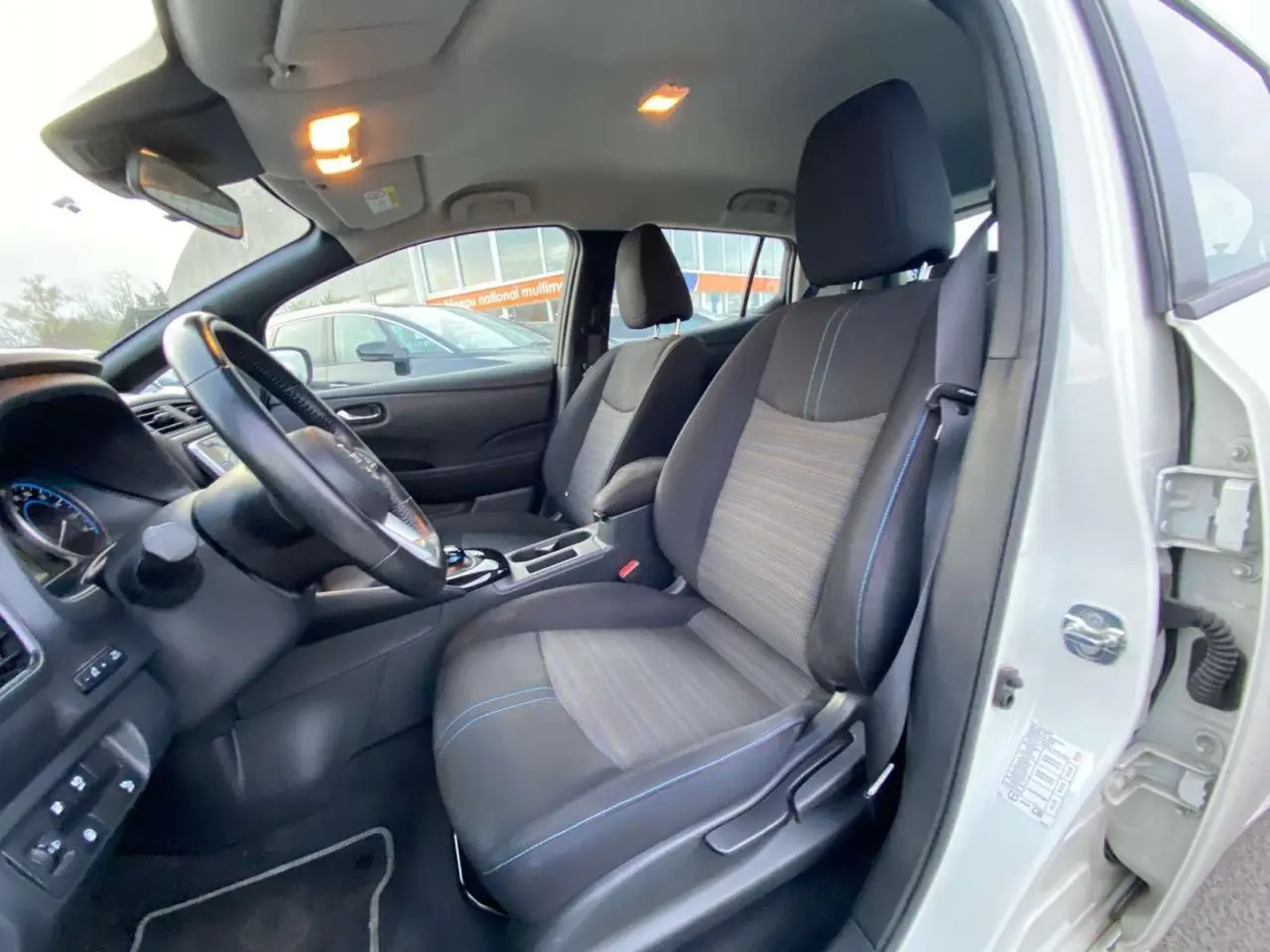 Vue intérieure côté conducteur d'une Nissan Leaf blanche 2020, sièges tissu noir et gris avec surpiqûres bleues.