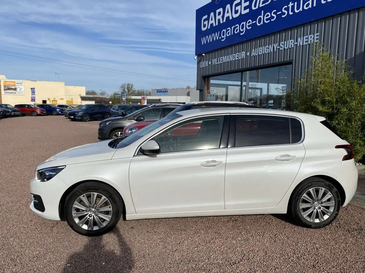 Profil côté gauche d'une Peugeot 308 blanche nacrée 2017 garée devant un garage avec vitres surteintées.
