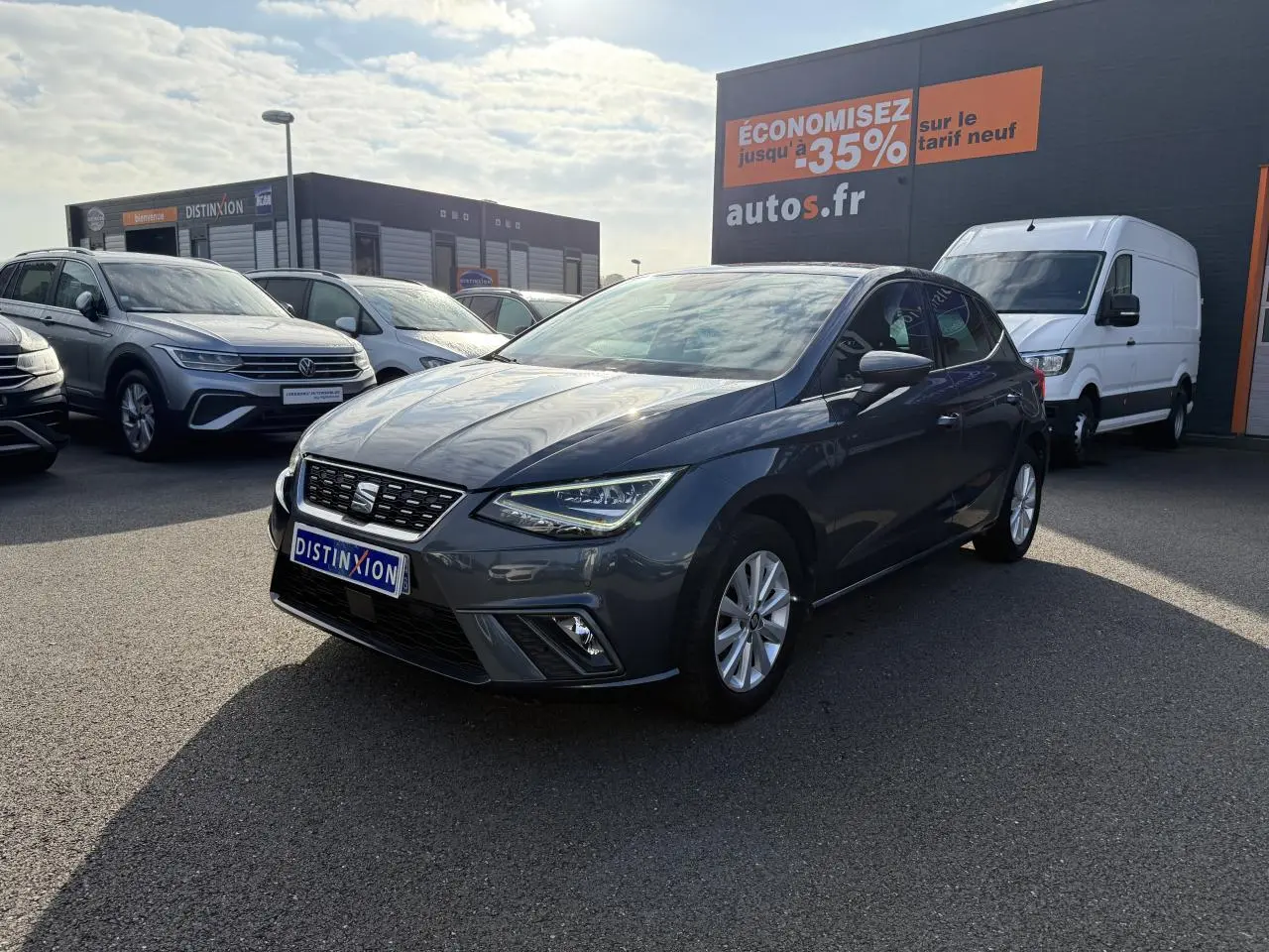SEAT Ibiza 1.0 EcoTSI 95 Xcellence gris Magnetic Tech vue 3/4 avant sur parking avec panneau promotionnel en arrière-plan