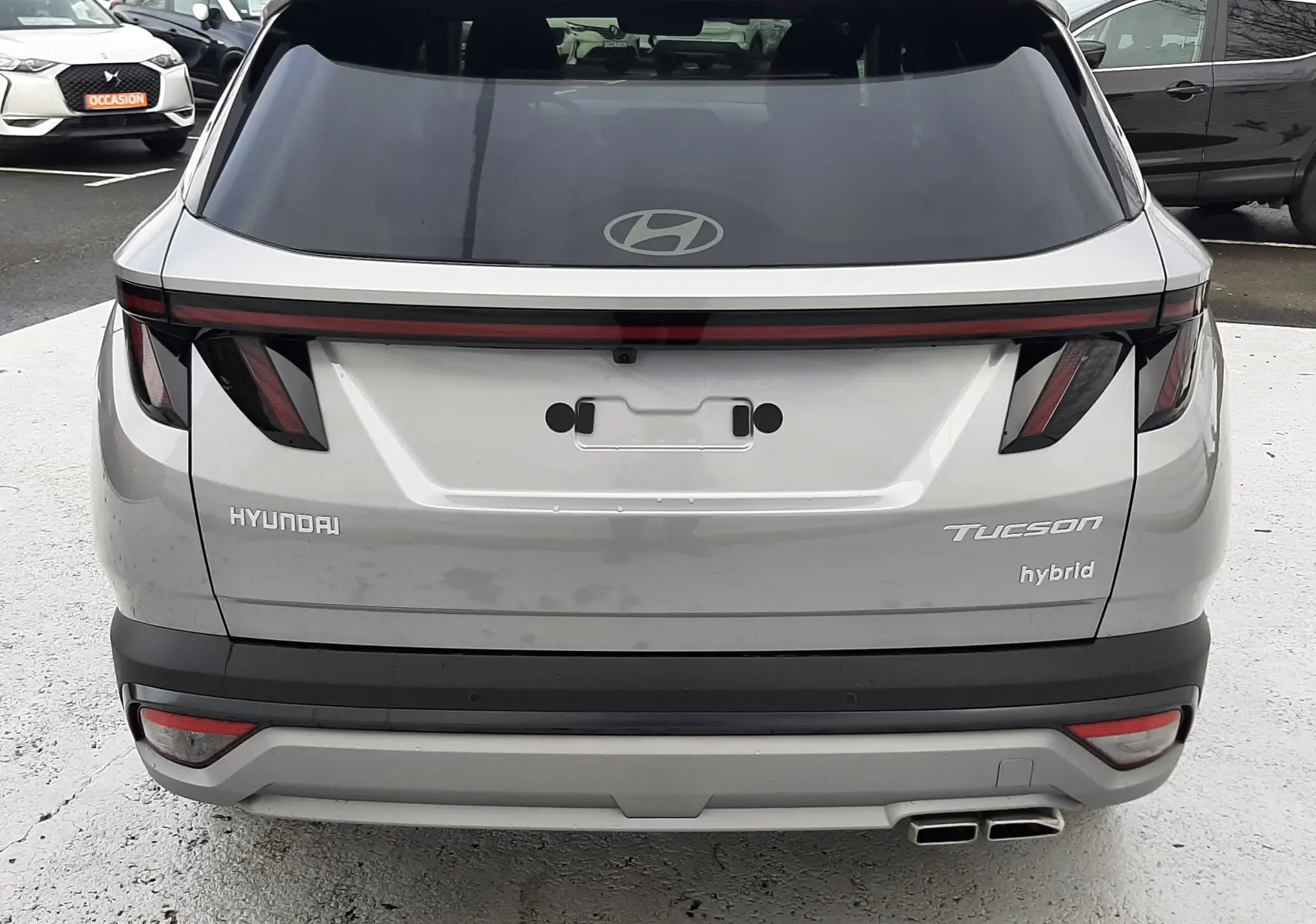 Vue arrière d’un Hyundai Tucson hybride gris Shimmering Silver avec feux arrière en bandeau lumineux et double sortie d’échappement.