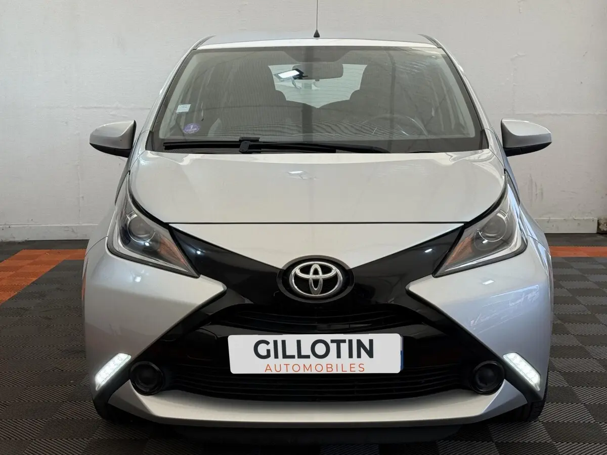 Vue avant frontale d'une Toyota Aygo gris clair avec calandre noire en forme de X et feux de jour LED allumés.