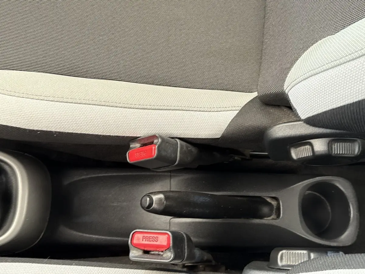 Vue rapprochée de la console centrale d'une Toyota Aygo gris clair, montrant le frein à main et les boucles de ceinture rouge.