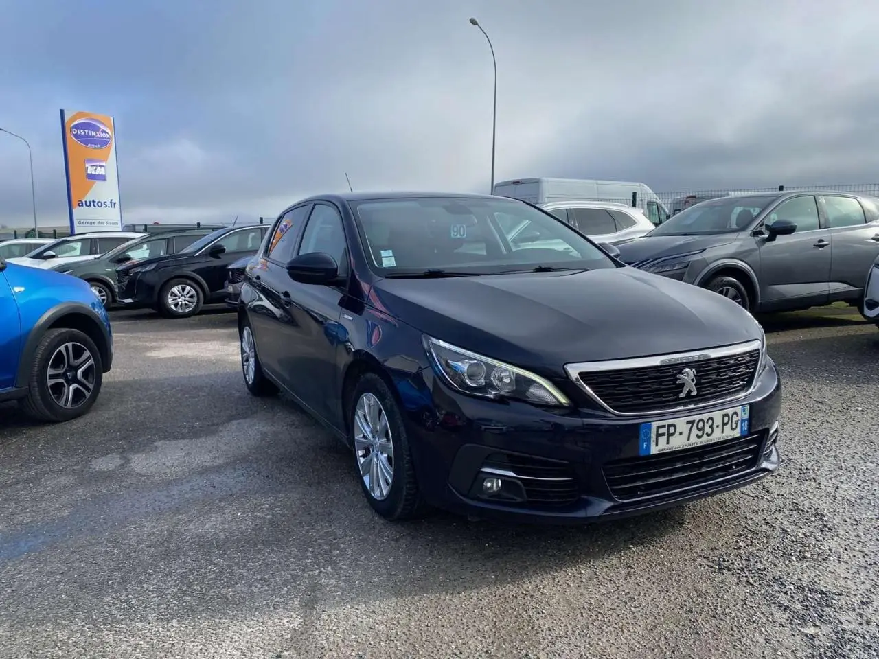 Peugeot 308 bleu foncé vue 3/4 avant droit sur parking avec calandre chromée et feux LED allumés.