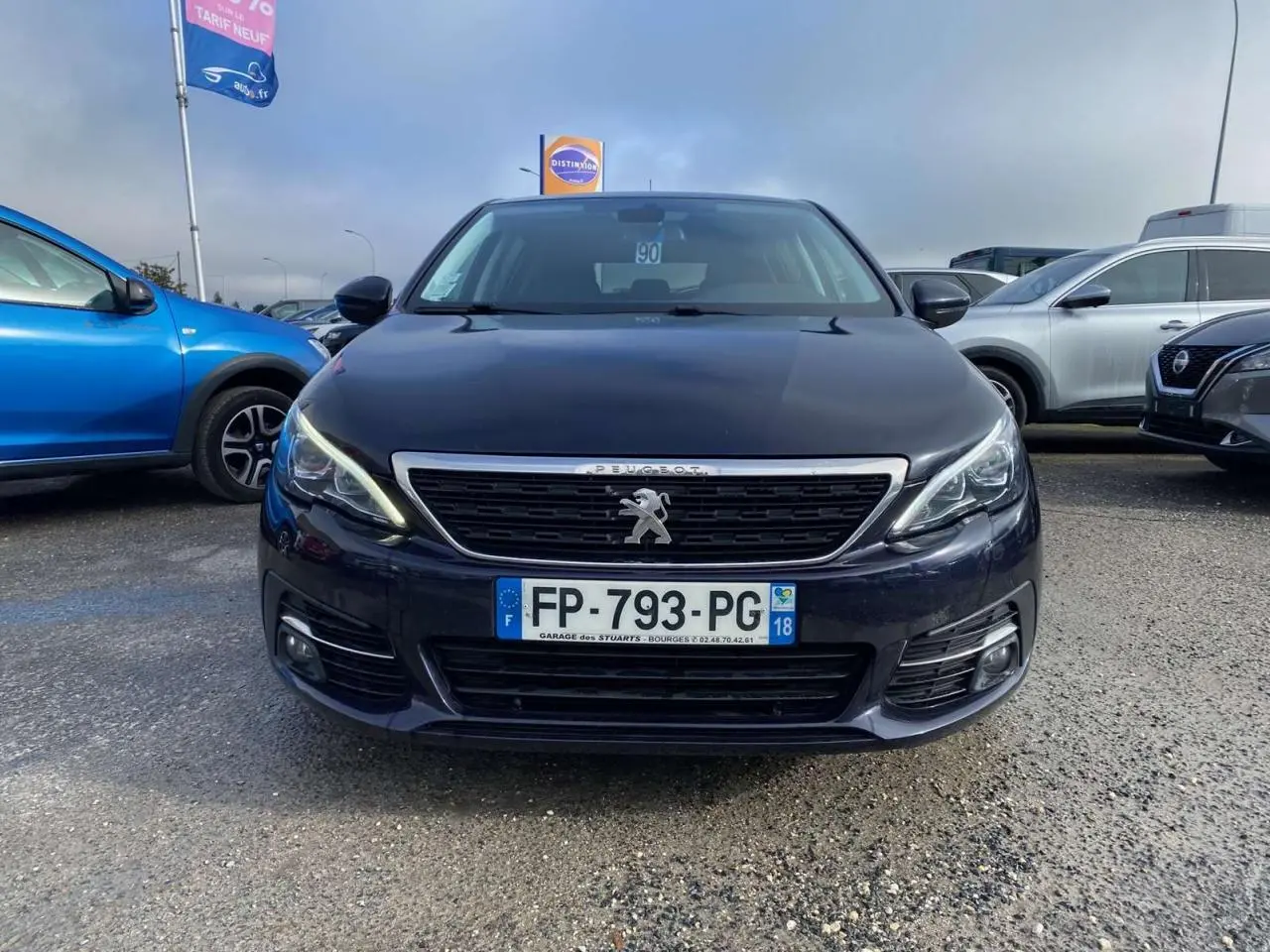 Peugeot 308 bleu foncé vue de face, avec calandre chromée et feux LED allumés, stationnée en extérieur.