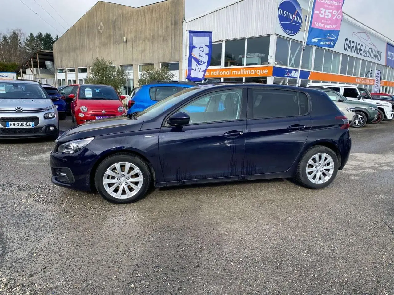 Peugeot 308 bleu foncé vue de profil côté gauche, garée devant un garage multimarque en extérieur.