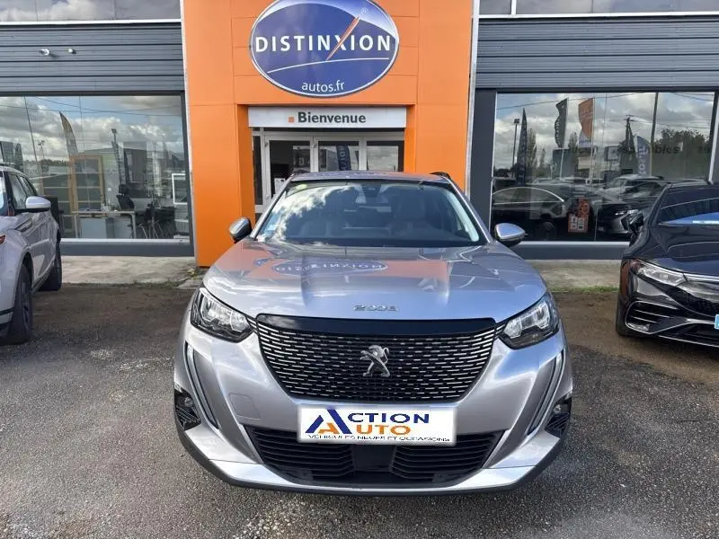 Peugeot 2008 gris Artense vue de face, calandre noire distinctive et feux LED allumés, devant un bâtiment orange.