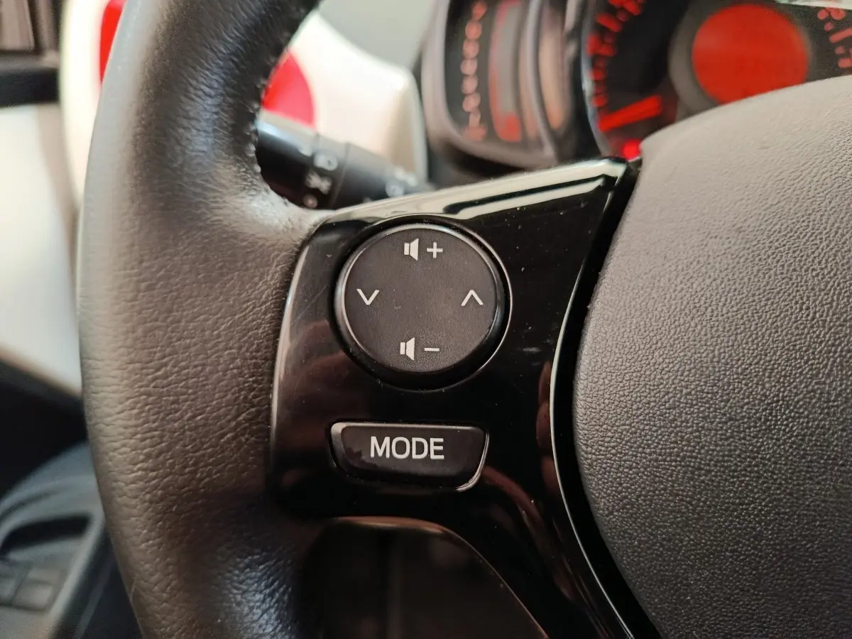 Gros plan sur les commandes audio au volant cuir noir de la Toyota Aygo 2016, avec tableau de bord rouge en arrière-plan.