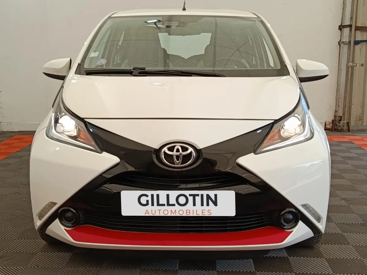 Vue avant d'une Toyota Aygo blanche avec calandre noire en X et détail rouge sous le pare-chocs, phares allumés.