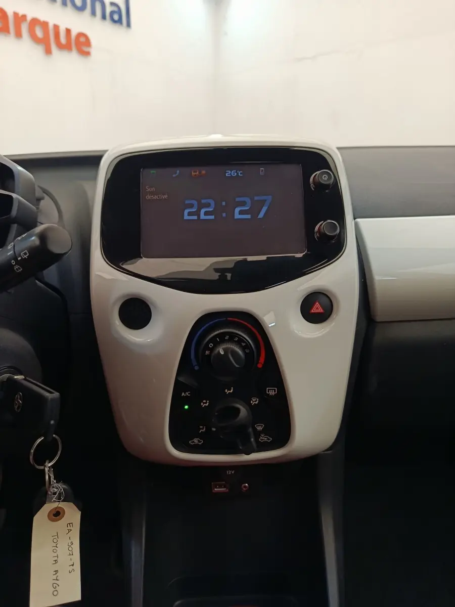 Vue rapprochée du tableau de bord blanc de la Toyota Aygo 2016, avec écran digital affichant l'heure et commandes de climatisation.