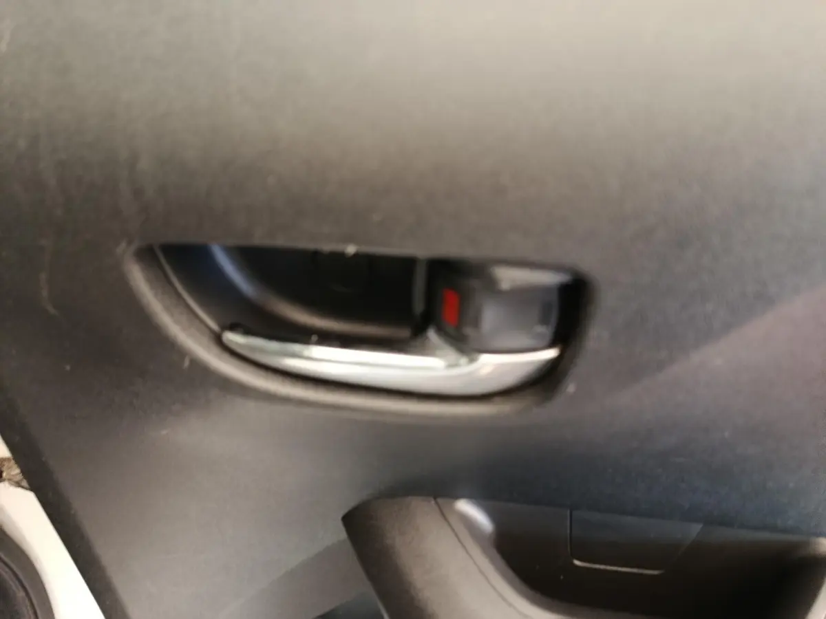 Poignée intérieure de porte avant gauche d'une Toyota Aygo blanche, finition 2016, avec insert chromé et bouton de verrouillage rouge.