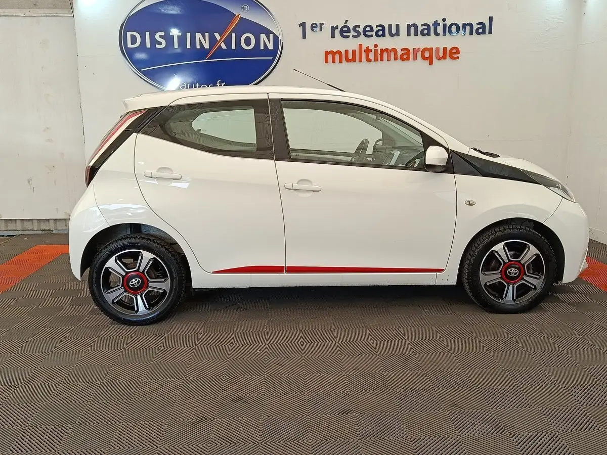Profil latéral droit de la Toyota Aygo blanche 2016 avec jantes noires et rouges et détails rouges sur les bas de caisse.