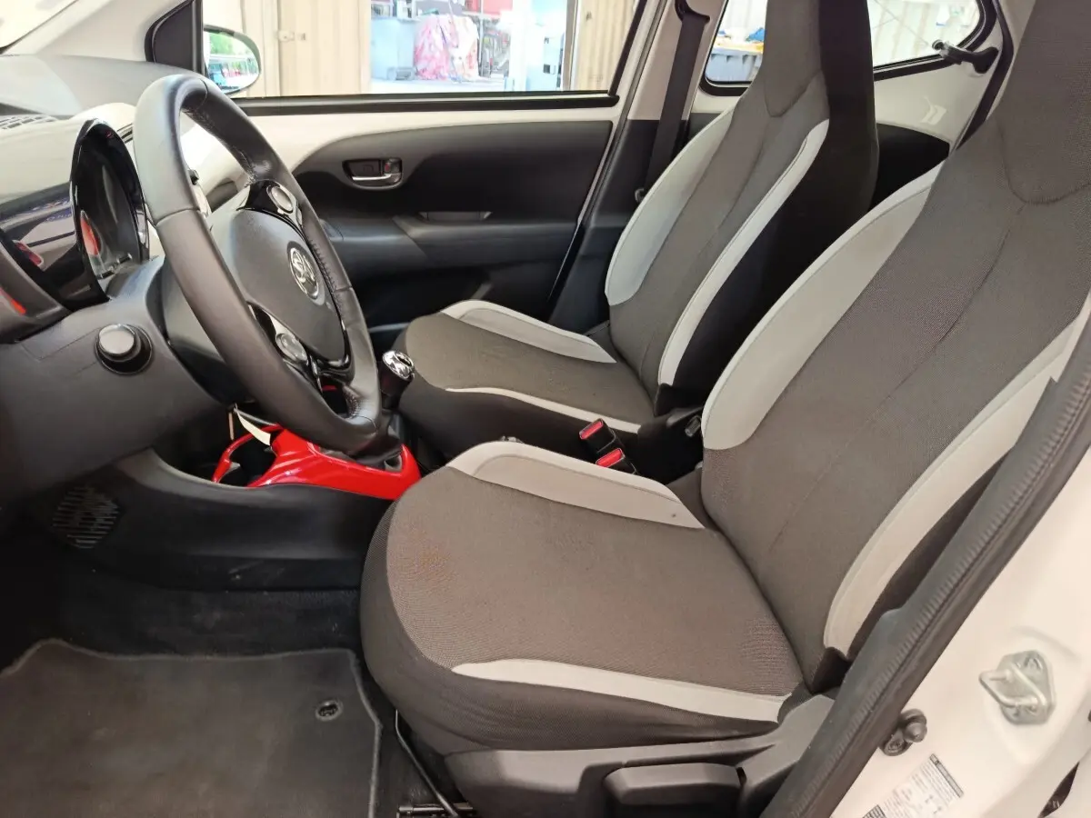 Vue intérieure côté conducteur de la Toyota Aygo blanche 2016, sièges gris avec console centrale rouge visible.