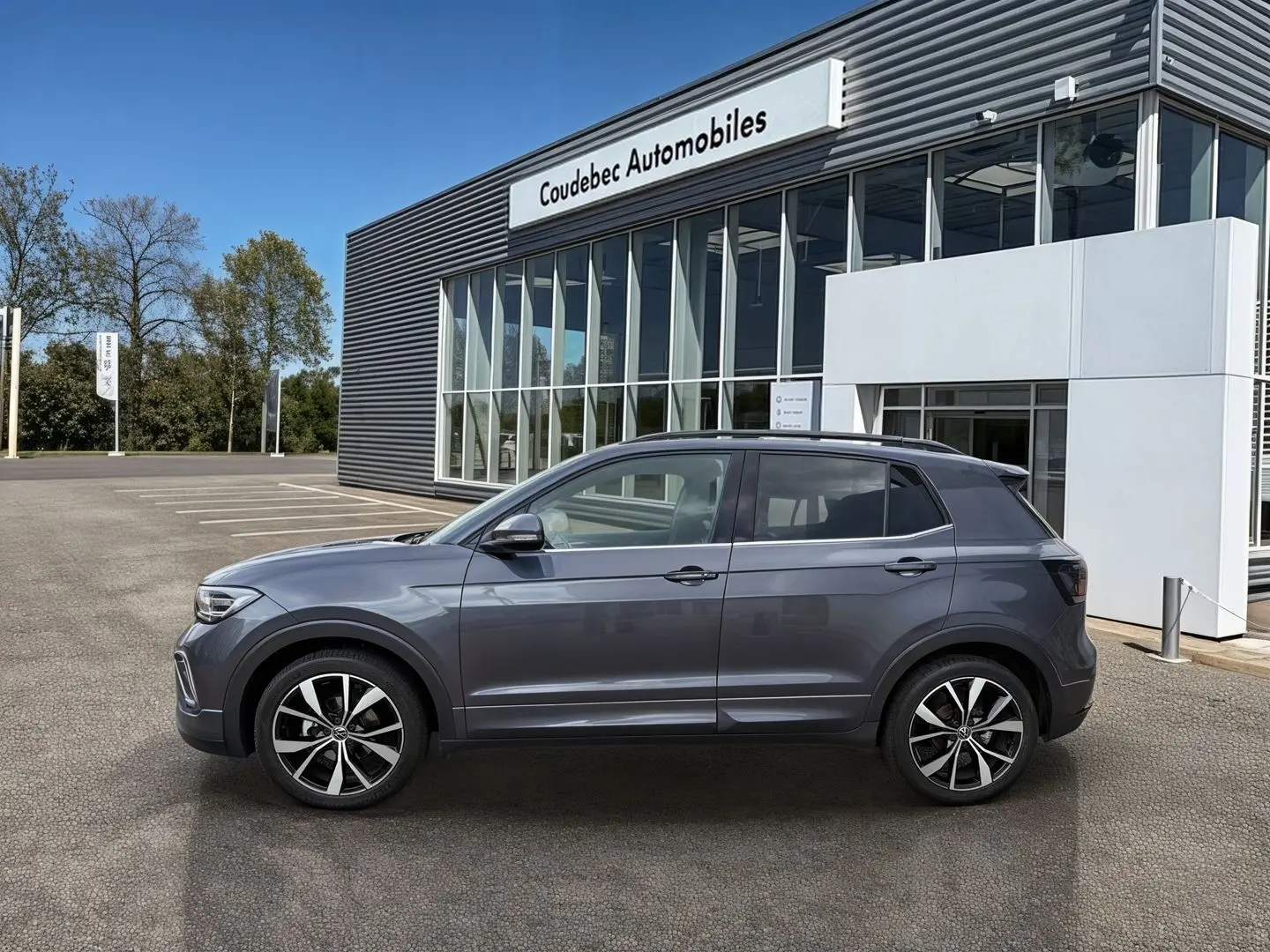 Vue de profil côté gauche d'un Volkswagen T-Cross gris 2025 avec jantes alliage 18 pouces devant un bâtiment commercial.