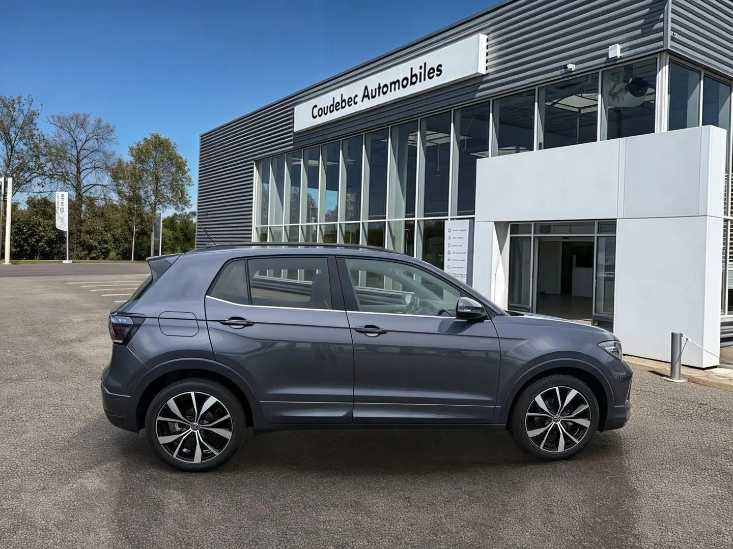Vue latérale droite d'un Volkswagen T-Cross gris 2025 avec jantes alliage noires et détails chromés, devant un concessionnaire.