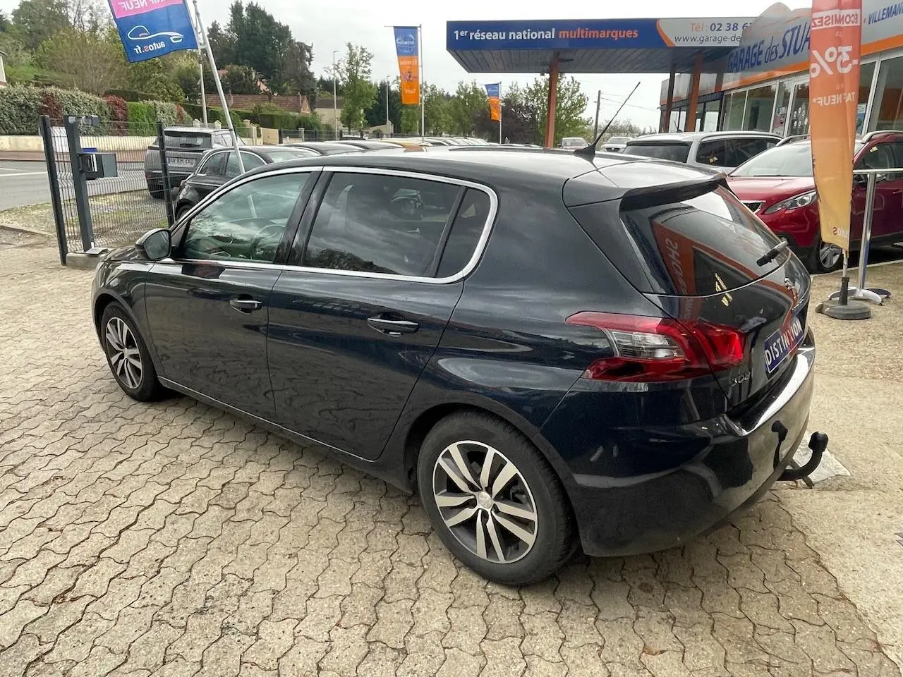 Vue 3/4 arrière droite d'une Peugeot 308 gris hurricane avec attelage de remorque, stationnée devant un garage.