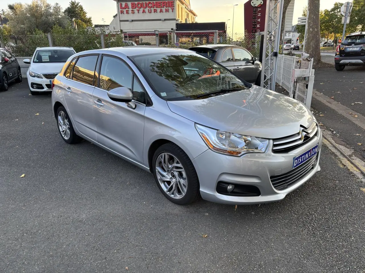 Citroën C4 gris clair vue 3/4 avant droit, stationnée devant un restaurant avec reflets métallisés.