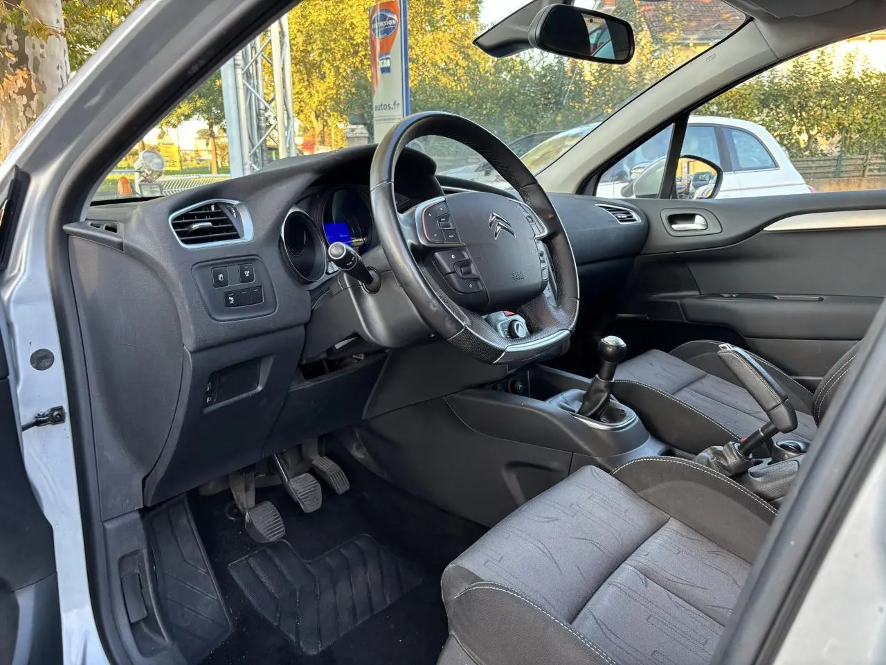 Intérieur côté conducteur d'une Citroën C4 gris clair 2012, volant multifonctions et boîte manuelle visibles.