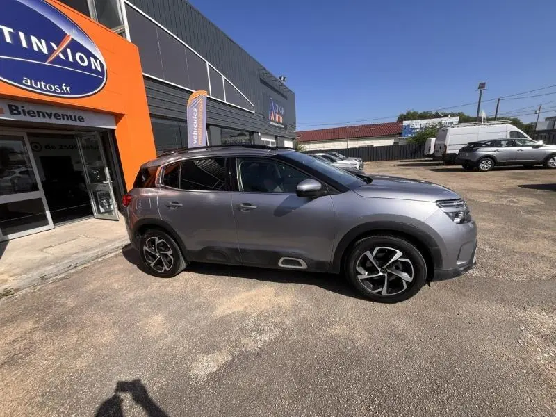 Vue de profil côté gauche d'un Citroën C5 Aircross gris métallisé garé devant un concessionnaire, avec jantes alliage distinctives.