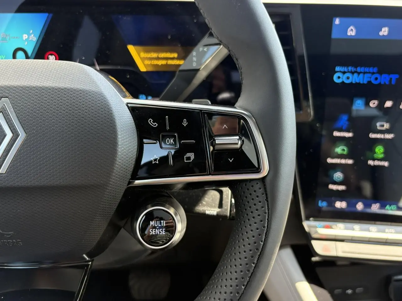Gros plan sur le volant noir perforé et les commandes multifonctions du Renault Espace VI hybride, écran multifonction en arrière-plan.