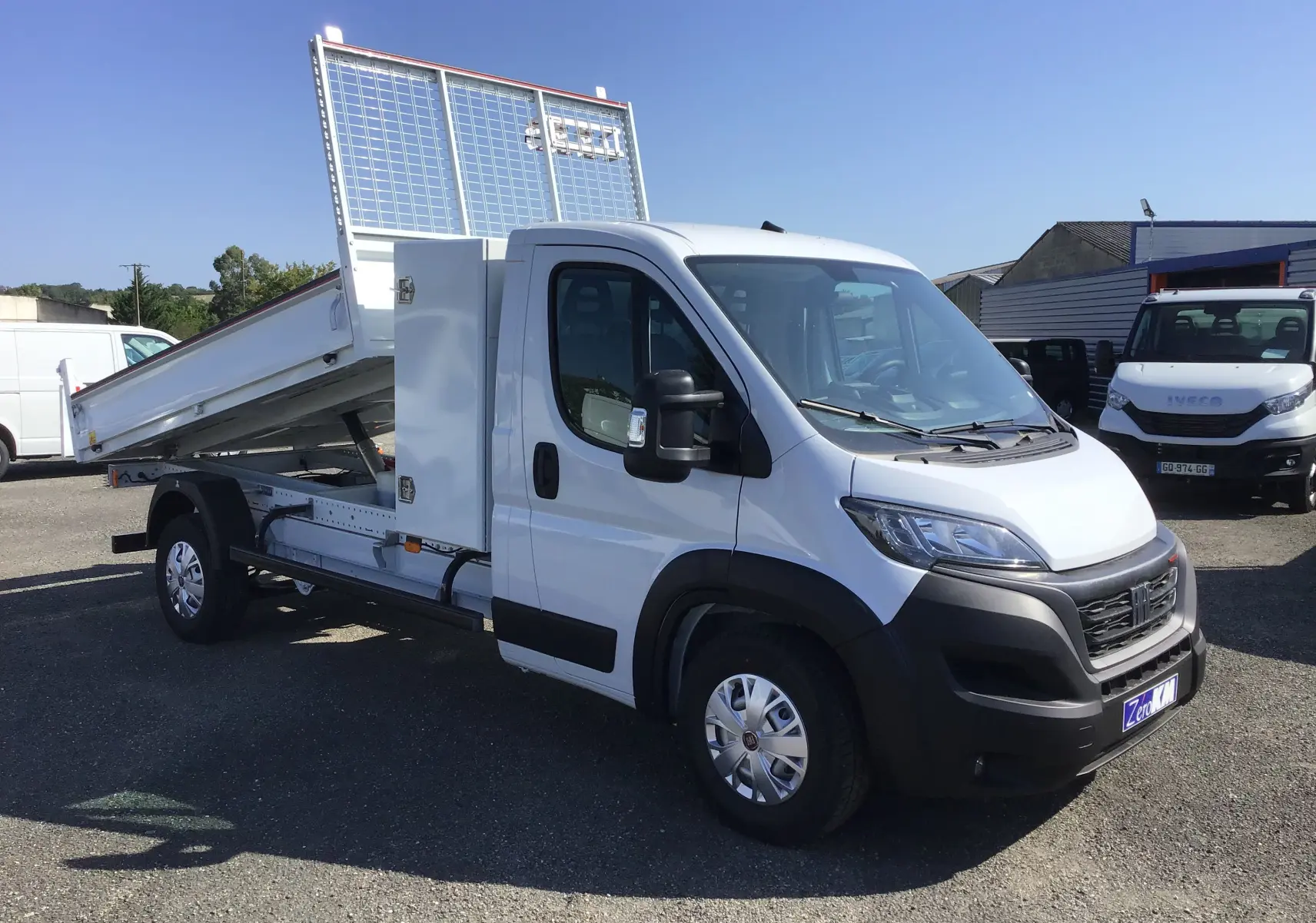 Vue 3/4 avant droite du FIAT Ducato Chassis Benne blanc 2023 avec benne acier relevée et coffre latéral.