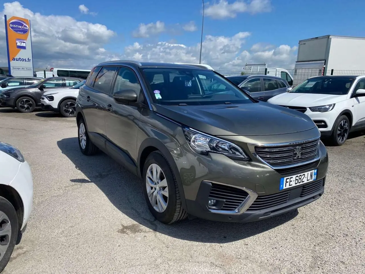 Peugeot 5008 gris clair vue 3/4 avant droit sur parking avec ciel partiellement nuageux.