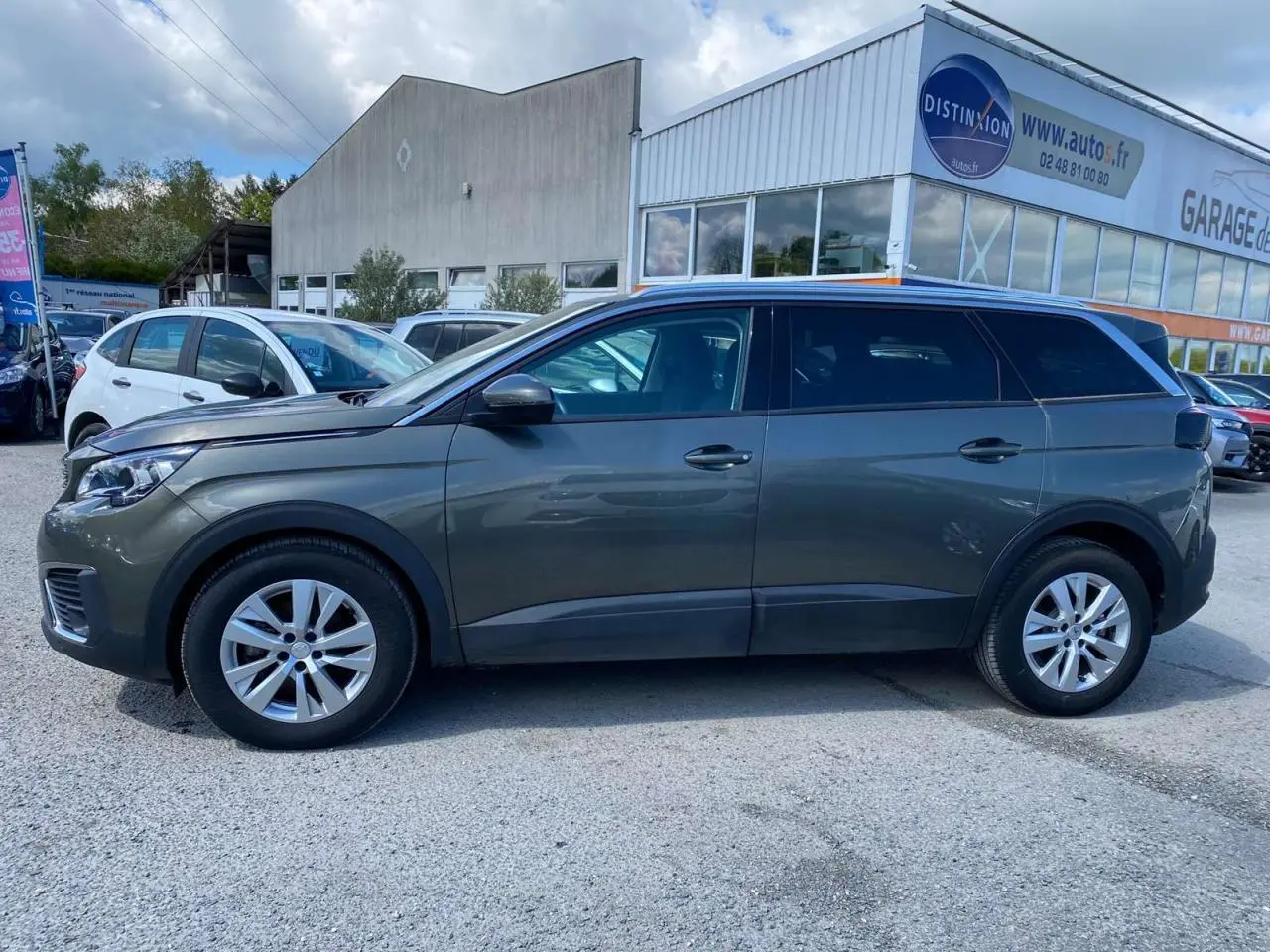 Profil côté gauche d'un Peugeot 5008 gris clair de 2019 garé devant un garage avec vitres surteintées.