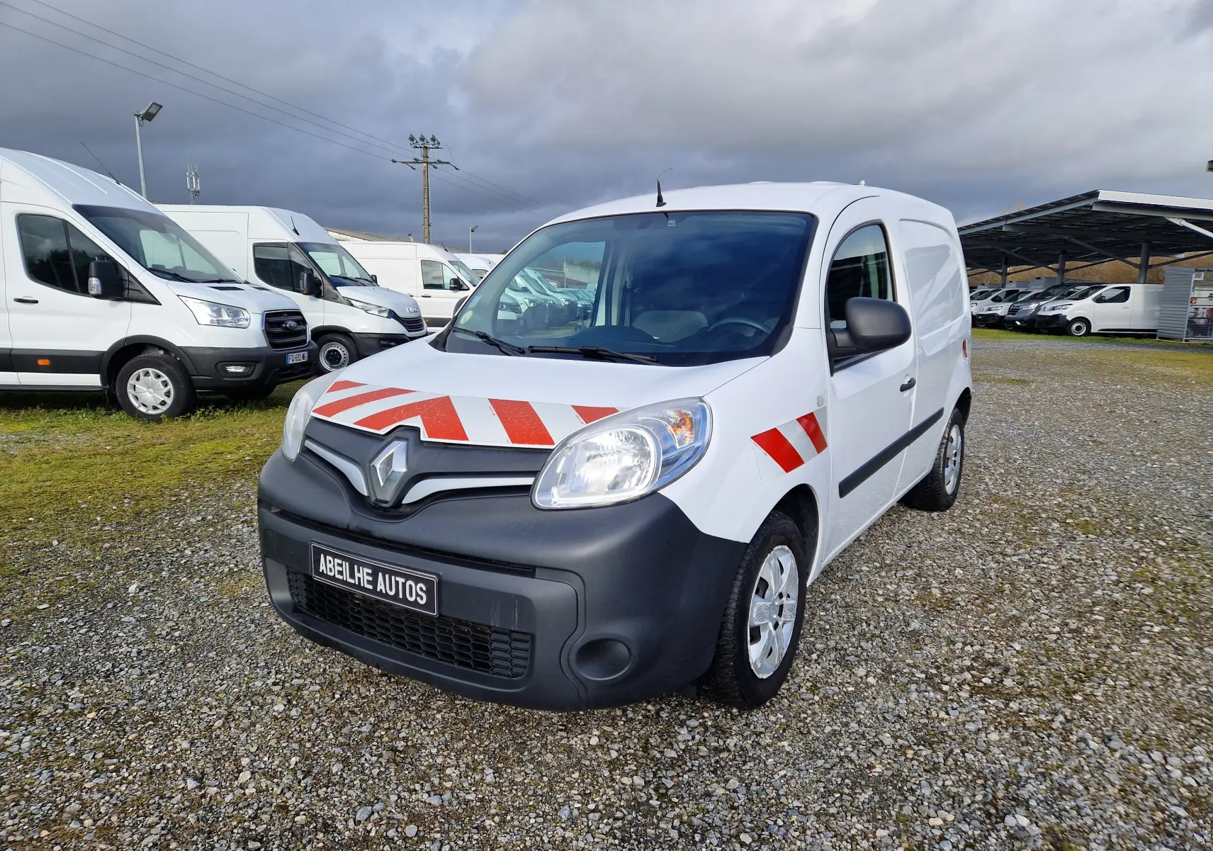 Vue 3/4 avant d’un Renault Kangoo blanc 2020 avec bandes rouges réfléchissantes sur capot et ailes, stationné sur gravier.