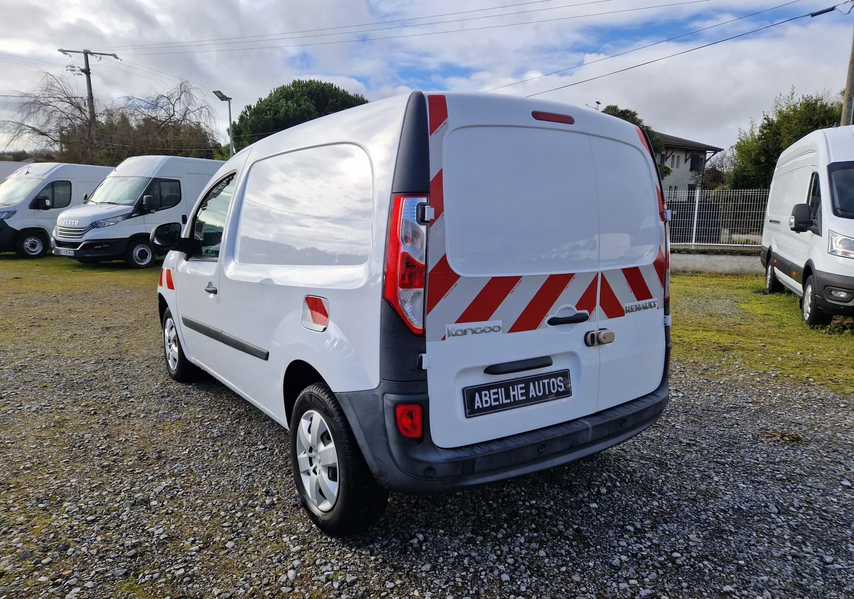 Vue 3/4 arrière droite d’un Renault Kangoo blanc avec bandes rouges réfléchissantes sur les portes arrière, stationné sur un sol gravillonné.