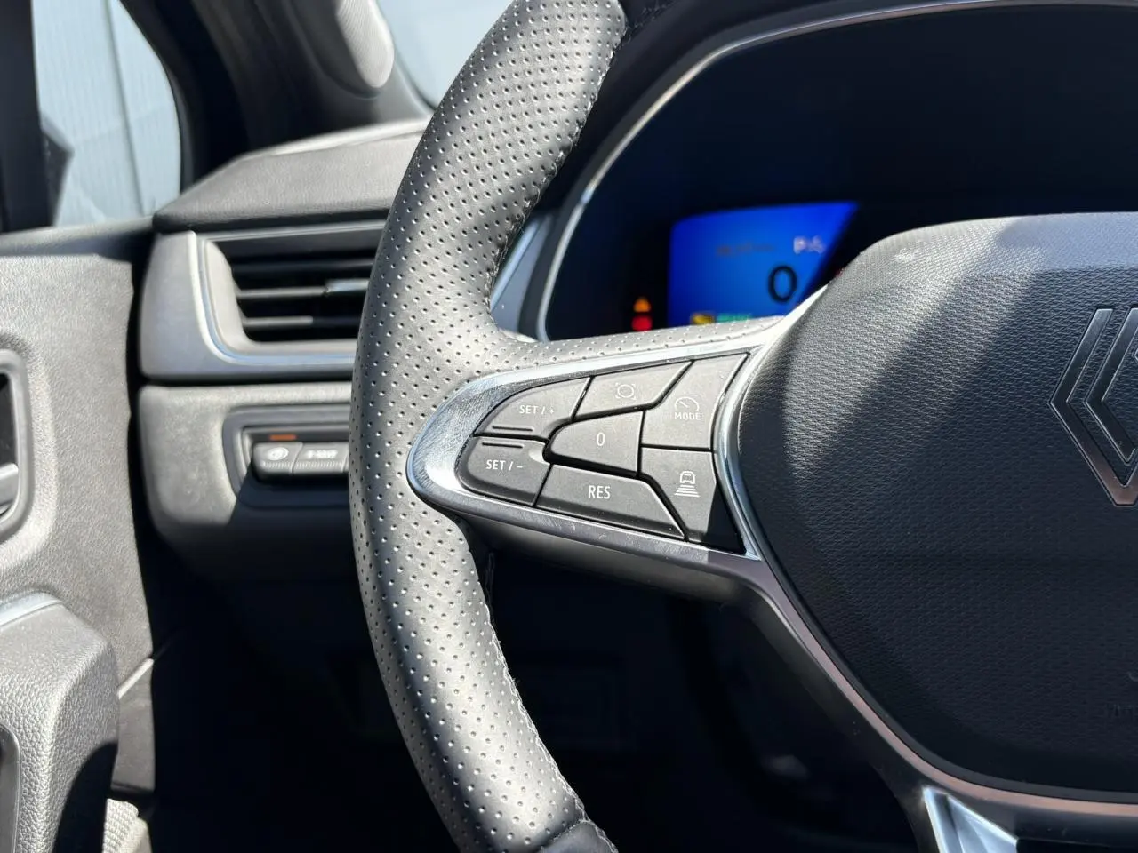 Gros plan sur le volant perforé de la Renault Symbioz noir étoilé, avec commandes intégrées et tableau de bord numérique bleu en arrière-plan.