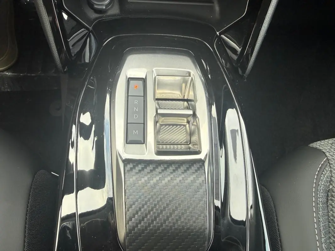 Vue plongeante sur la console centrale noire brillante de la Peugeot 208 Hybrid 145 avec levier de vitesses électronique et finition carbone.