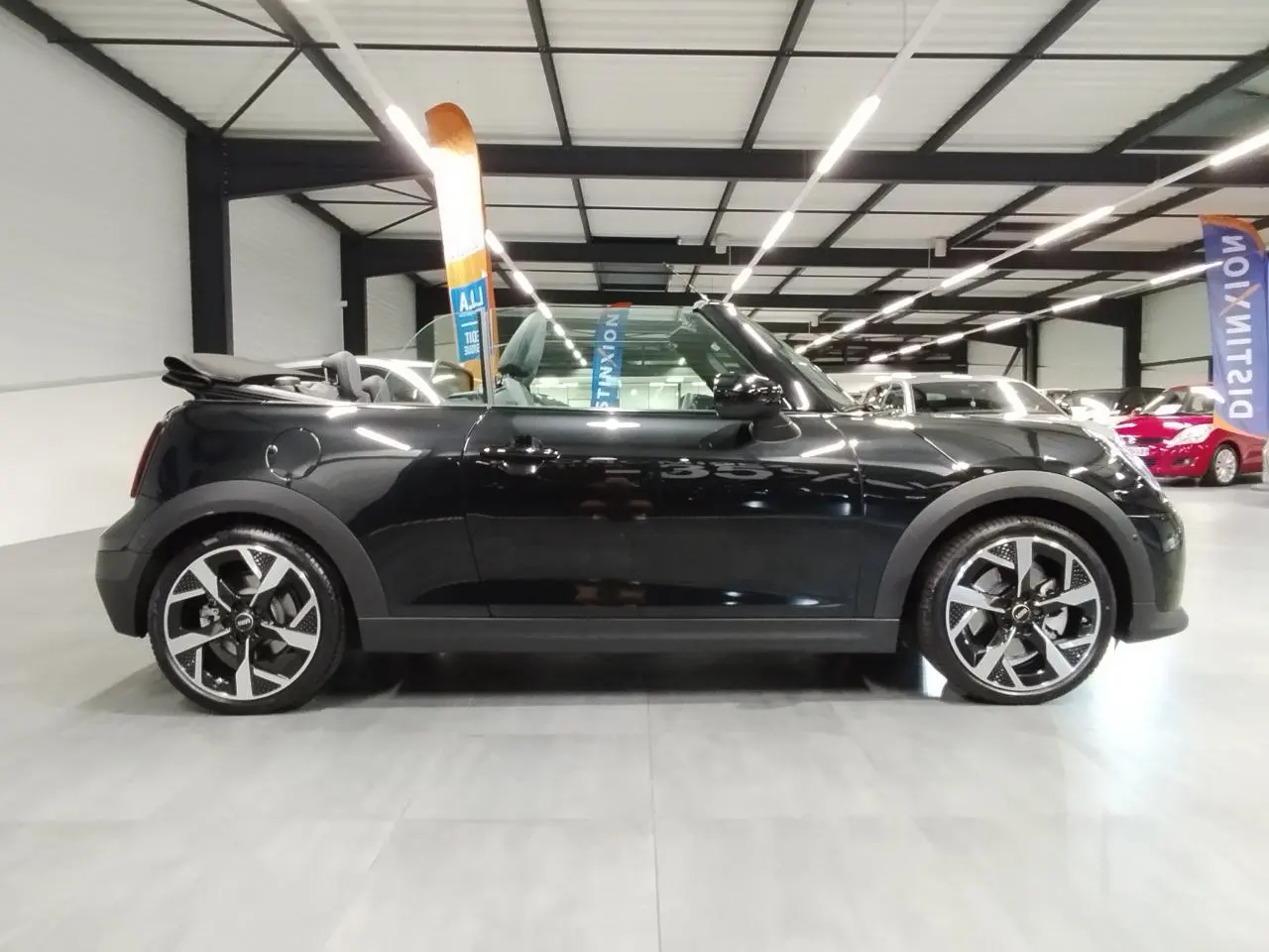Profil droit d'une MINI Cabriolet noire 2025 avec capote ouverte et jantes 16 pouces 4-Square Spoke en intérieur showroom.