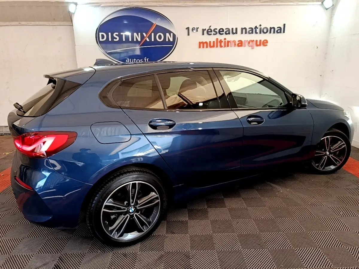 BMW Série 1 118i Edition Sport bleu vue de profil droit en intérieur, avec jantes alliage et feux arrière LED allumés.